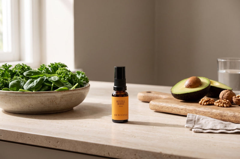 Spray B12 Cell Boost de Biologyst, flacon de 20 ml, sur un plan de travail en travertin, entre un bol d'épinards et de chou kale et une planche avec un avocat et des noix, à la lumière du matin